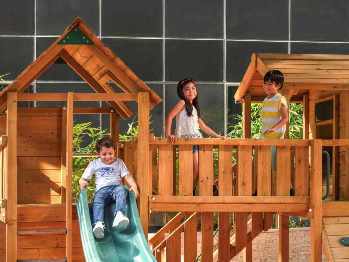 Grand Mercure Dubai City Hotel Exterior photo The photo shows three children playing on a wooden playground structure. There is a slide on which a young boy is sitting, preparing to slide down. Two other children, a boy and a girl, are standing on a raised platform of the playground. The backgro