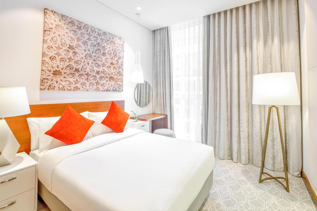 Grand Mercure Dubai City Hotel Exterior photo The photo shows a modern hotel room featuring a comfortable queen-sized bed with white linens and orange accent pillows. There is a decorative wall art piece above the bed, adding a stylish touch to the room. A round mirror is mounted on the wall abo