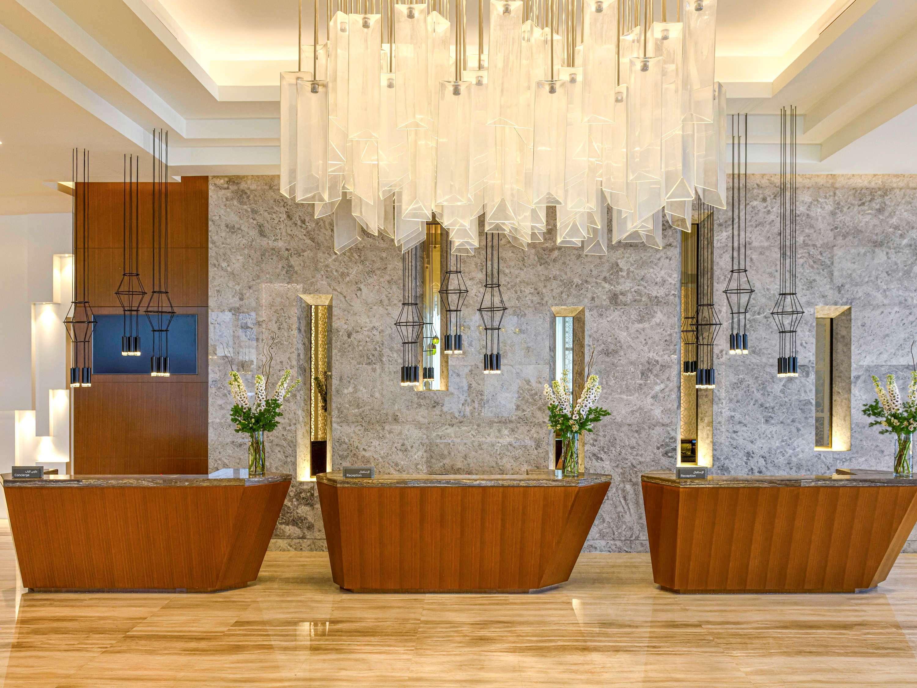 Grand Mercure Dubai City Hotel Exterior photo The photo displays a modern hotel reception area characterized by a sleek and stylish design. In the foreground, there are three wooden reception desks that have a smooth, curved shape. Behind the desks, the wall features a mix of marble and decorati