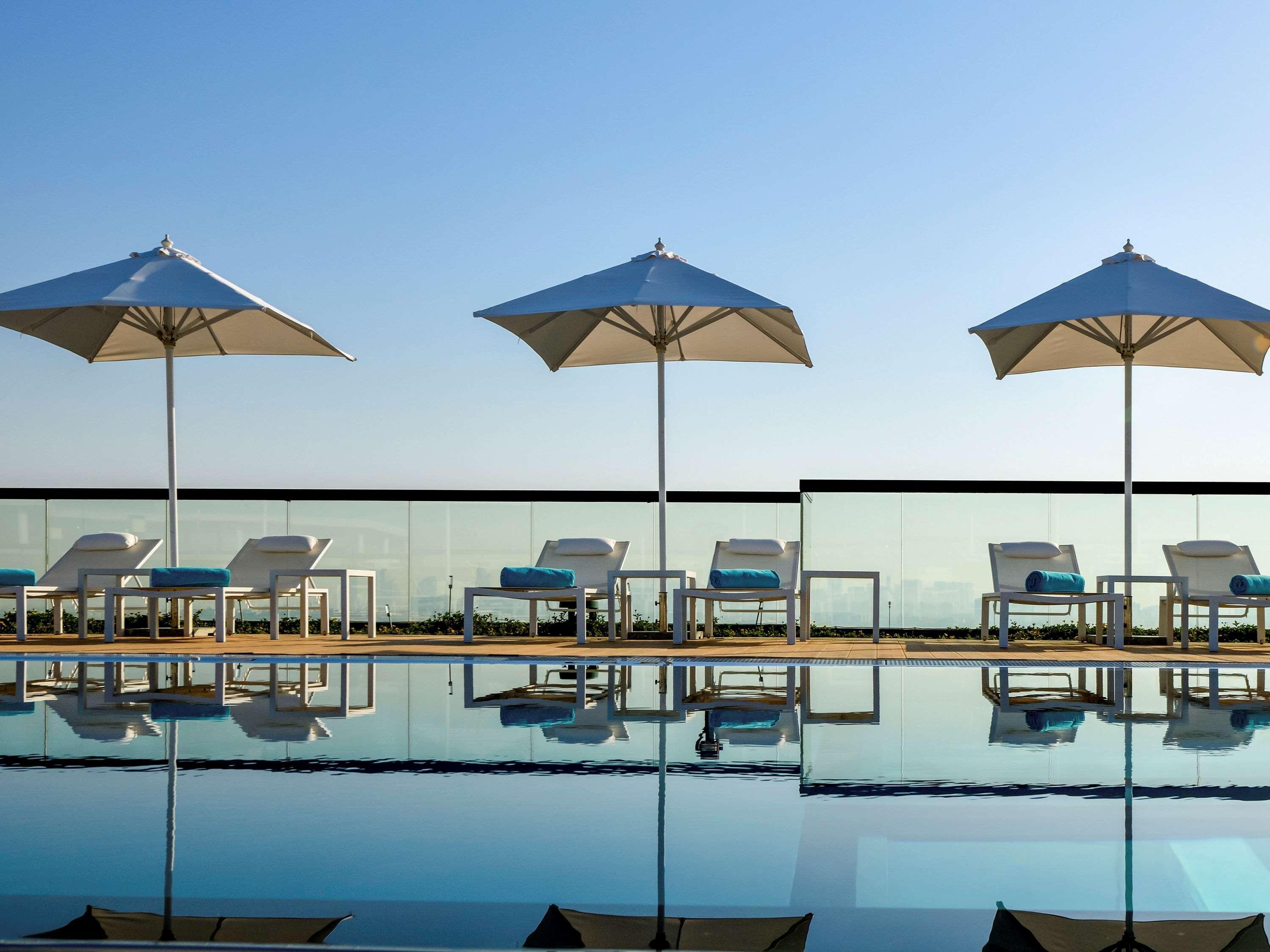 Grand Mercure Dubai City Hotel Exterior photo The photo shows a serene poolside scene featuring a series of white lounge chairs with blue cushions positioned alongside a swimming pool. Each chair is shaded by a large, white umbrella. The pool reflects the bright blue sky above, creating a tranqu