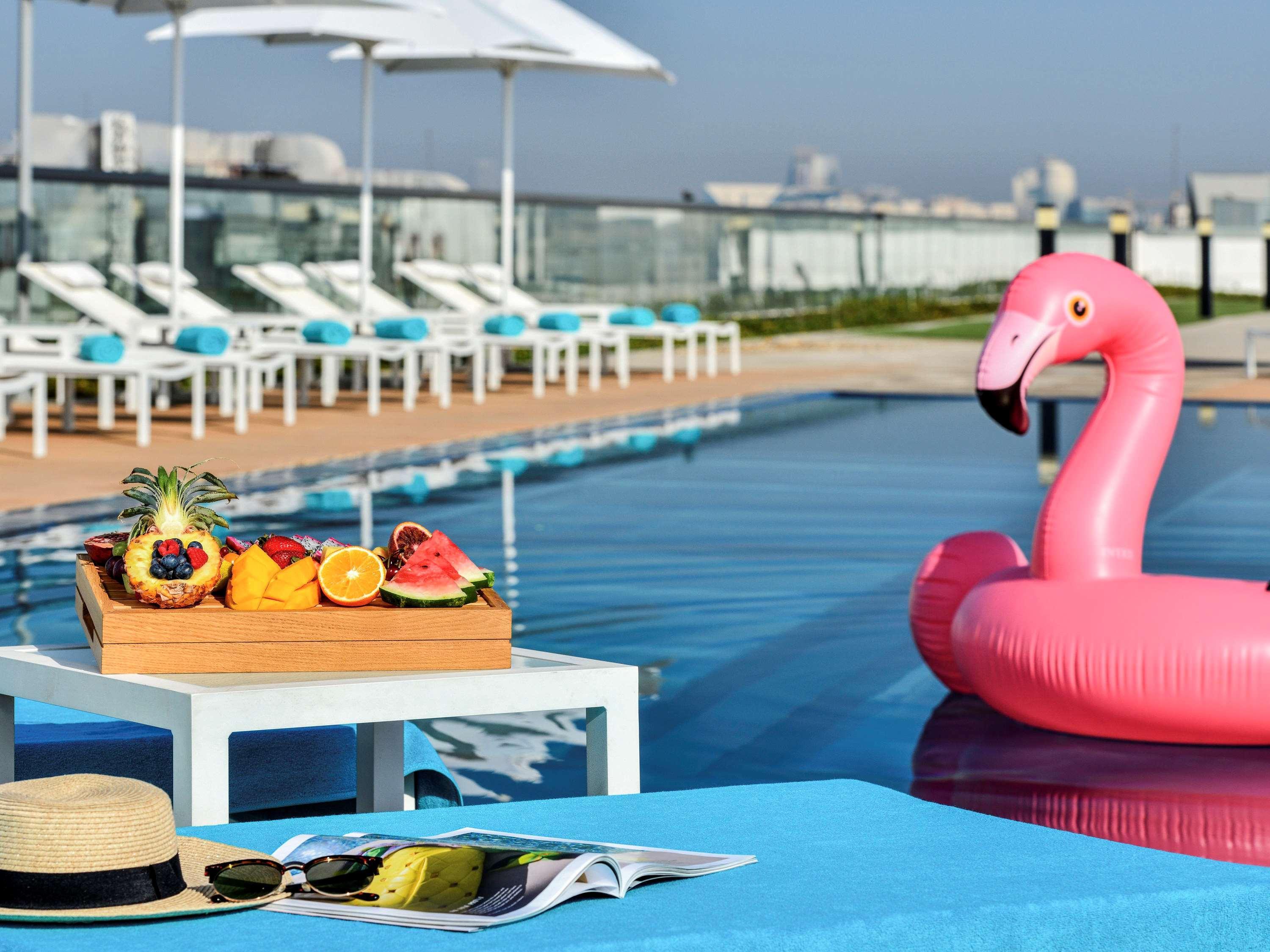 Grand Mercure Dubai City Hotel Exterior photo The photo depicts a relaxing poolside scene. In the foreground, there's a small white table with a wooden tray filled with a variety of colorful fruits, including berries, a slice of melon, and citrus fruits. Nearby, there's a beach hat and a magazin