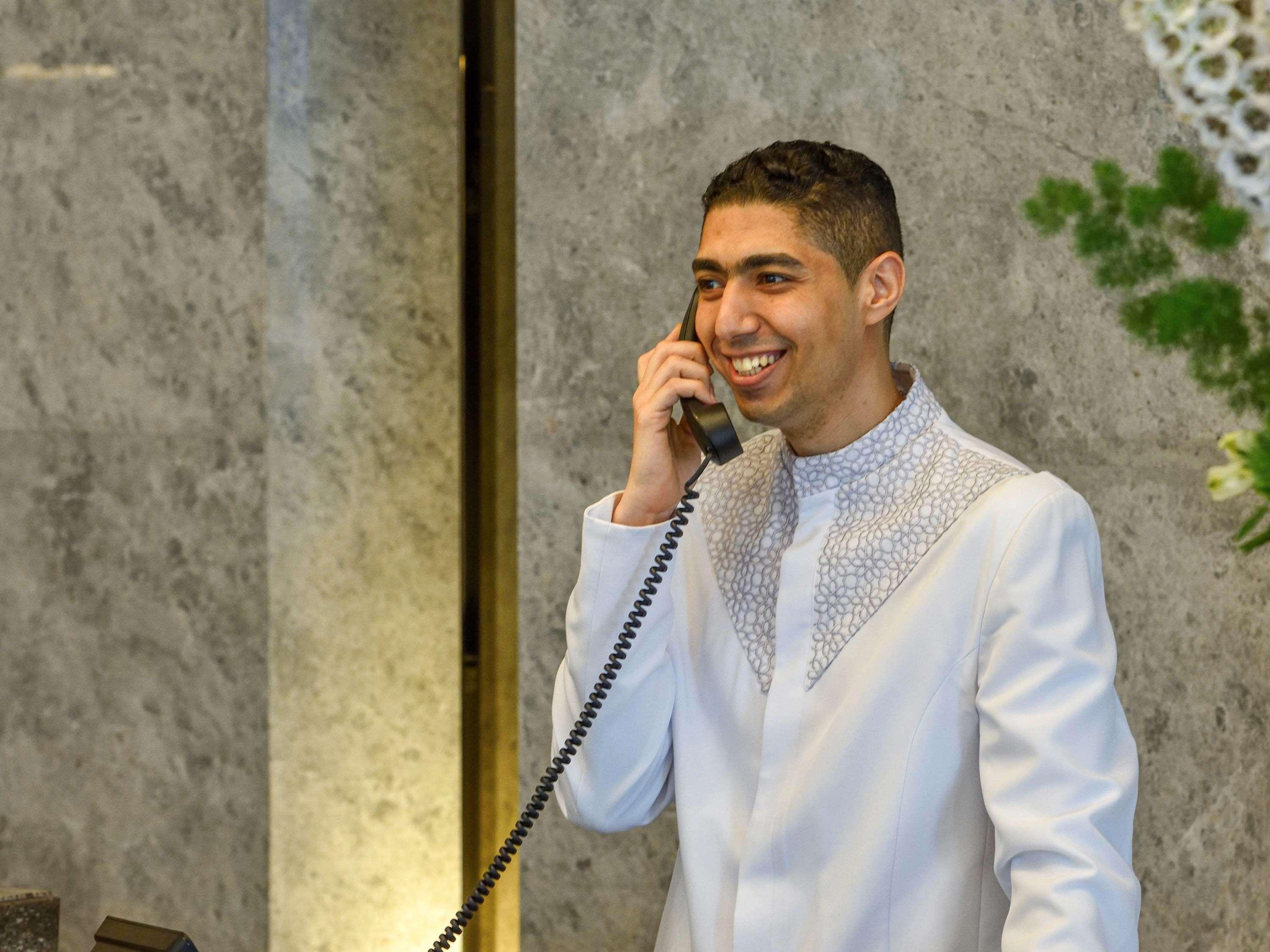 Grand Mercure Dubai City Hotel Exterior photo The photo shows a young man dressed in a formal white outfit with decorative details around the collar. He is smiling and is speaking on a vintage-style telephone. The background appears to have a marble texture, and there are some plants or floral a