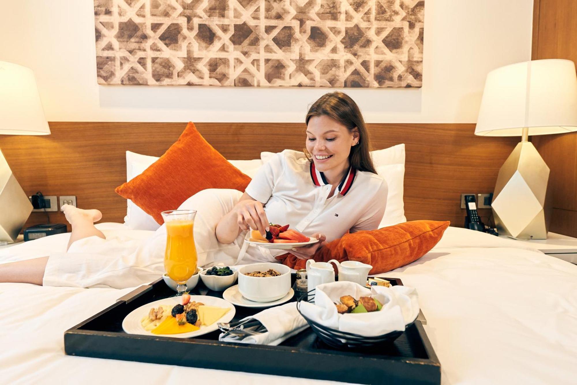 Grand Mercure Dubai City Hotel Exterior photo The photo shows a young woman sitting on a bed, enjoying a breakfast tray in front of her. The tray is filled with an assortment of food, including a bowl of fruit, a dish of cereal or yogurt, and some pastries. Next to the food, there is a glass of 