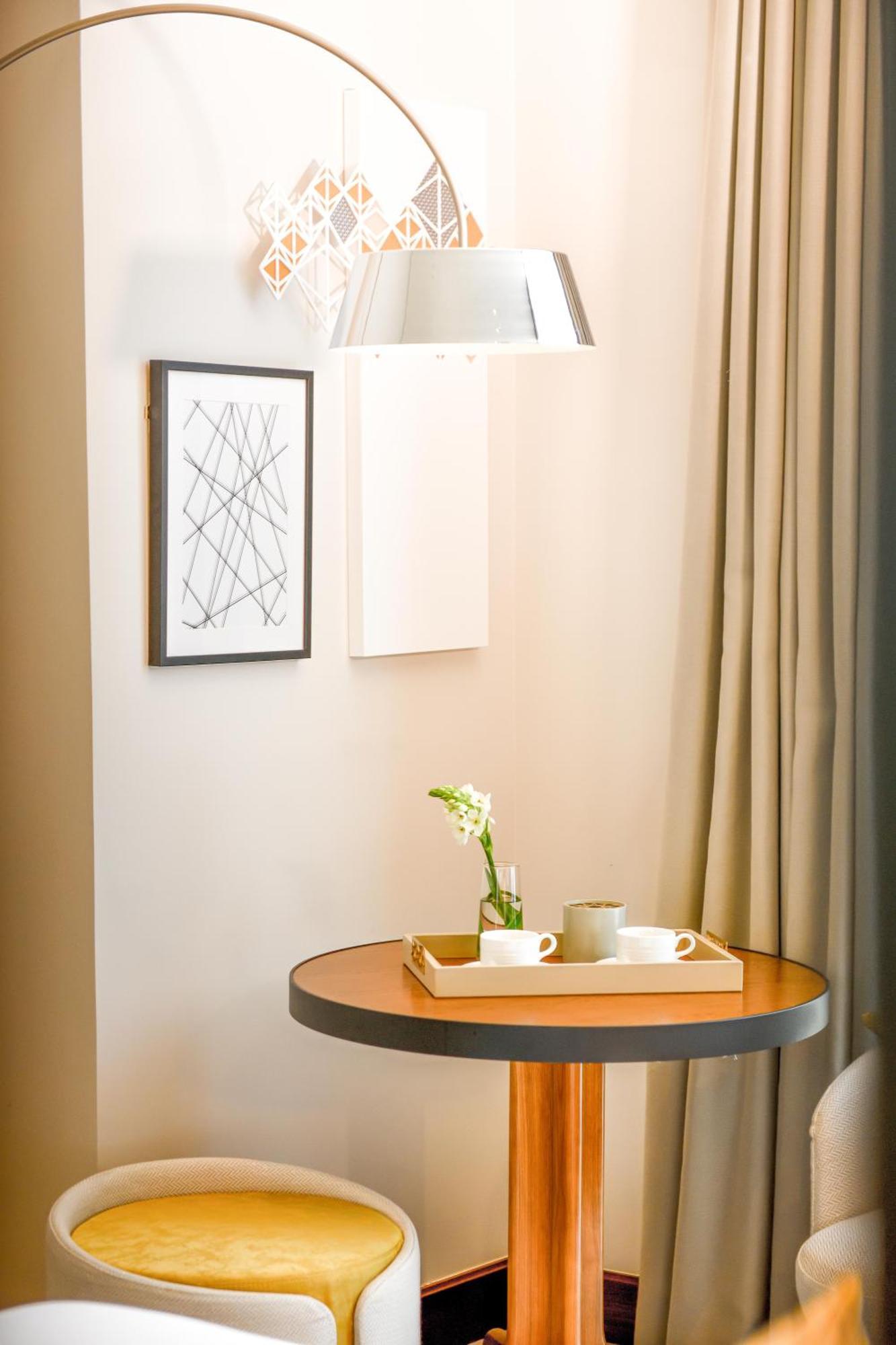 Grand Mercure Dubai City Hotel Exterior photo The photo shows a small table with a round wooden surface and a light-colored tray on top. The tray holds two white cups, a light green mug, and a small vase with a flower. In the background, there is a piece of framed artwork featuring abstract, int