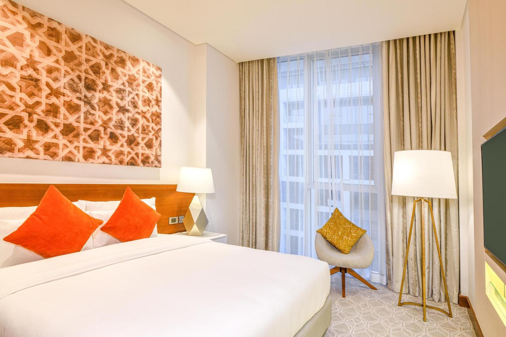 Grand Mercure Dubai City Hotel Exterior photo The photo shows a modern hotel room with a cozy and elegant design. There is a large bed with crisp white linens, accompanied by decorative cushions in shades of orange. To the side, there are two bedside lamps providing soft lighting. A colorful pie