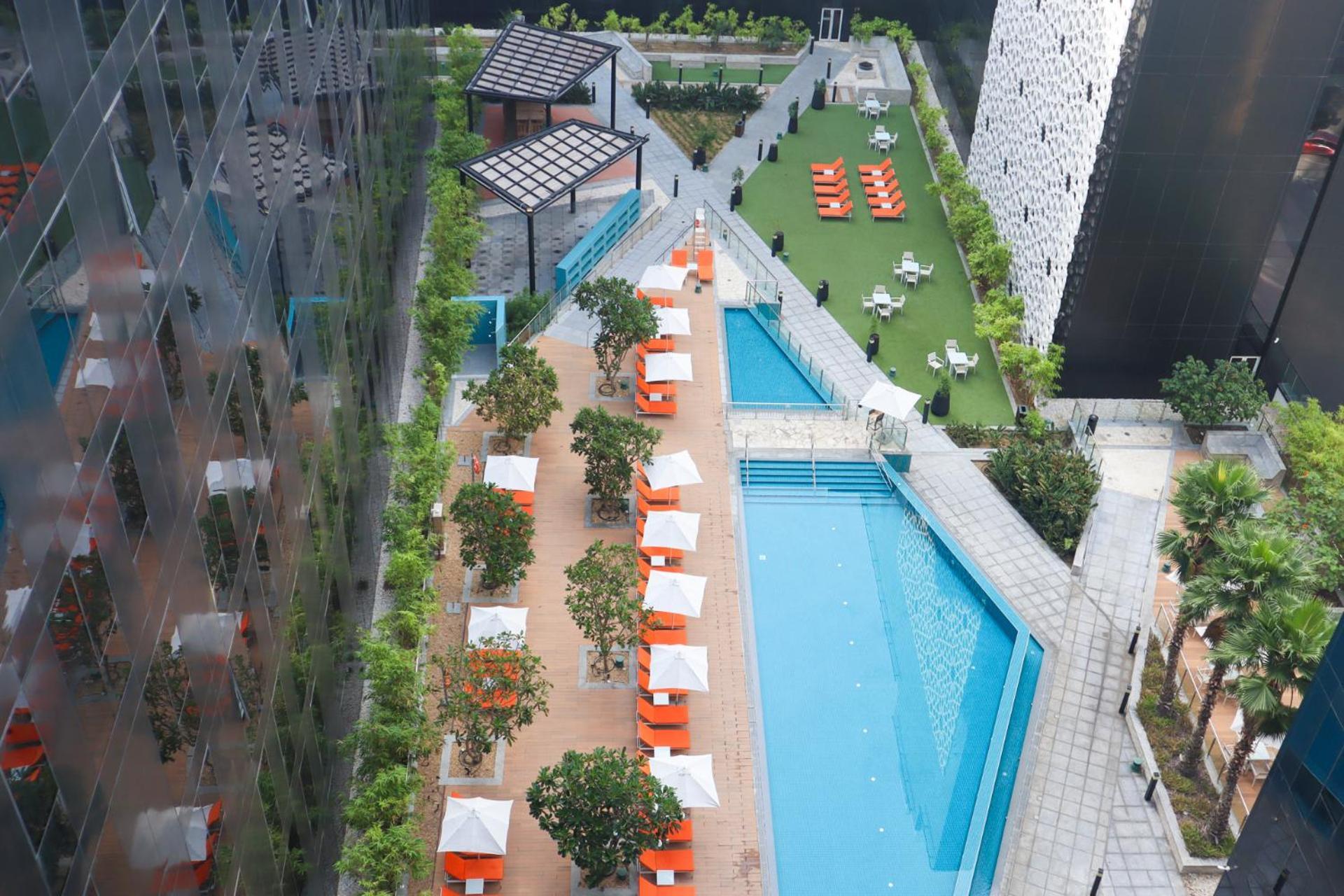 Grand Mercure Dubai City Hotel Exterior photo The photo depicts a rooftop pool area surrounded by greenery and lounging spaces. On one side, there is a large swimming pool, and adjacent to it, several orange lounge chairs and shaded areas with white canopies are set up for relaxation. The landsc