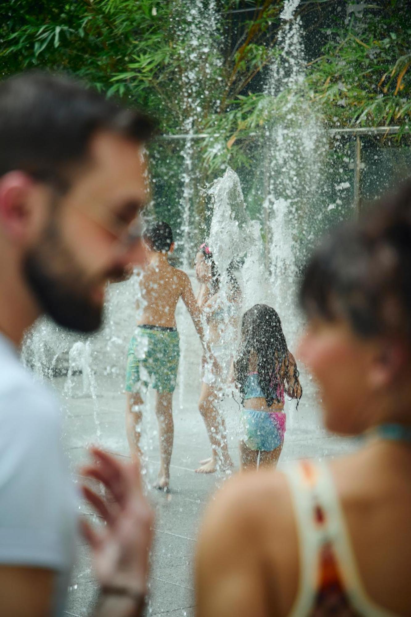 Grand Mercure Dubai City Hotel Exterior photo The photo shows a vibrant scene at a water fountain. In the foreground, two people are partially visible, one with a beard and sunglasses and the other with long hair. In the background, children are playing in the fountain, enjoying the water as it 