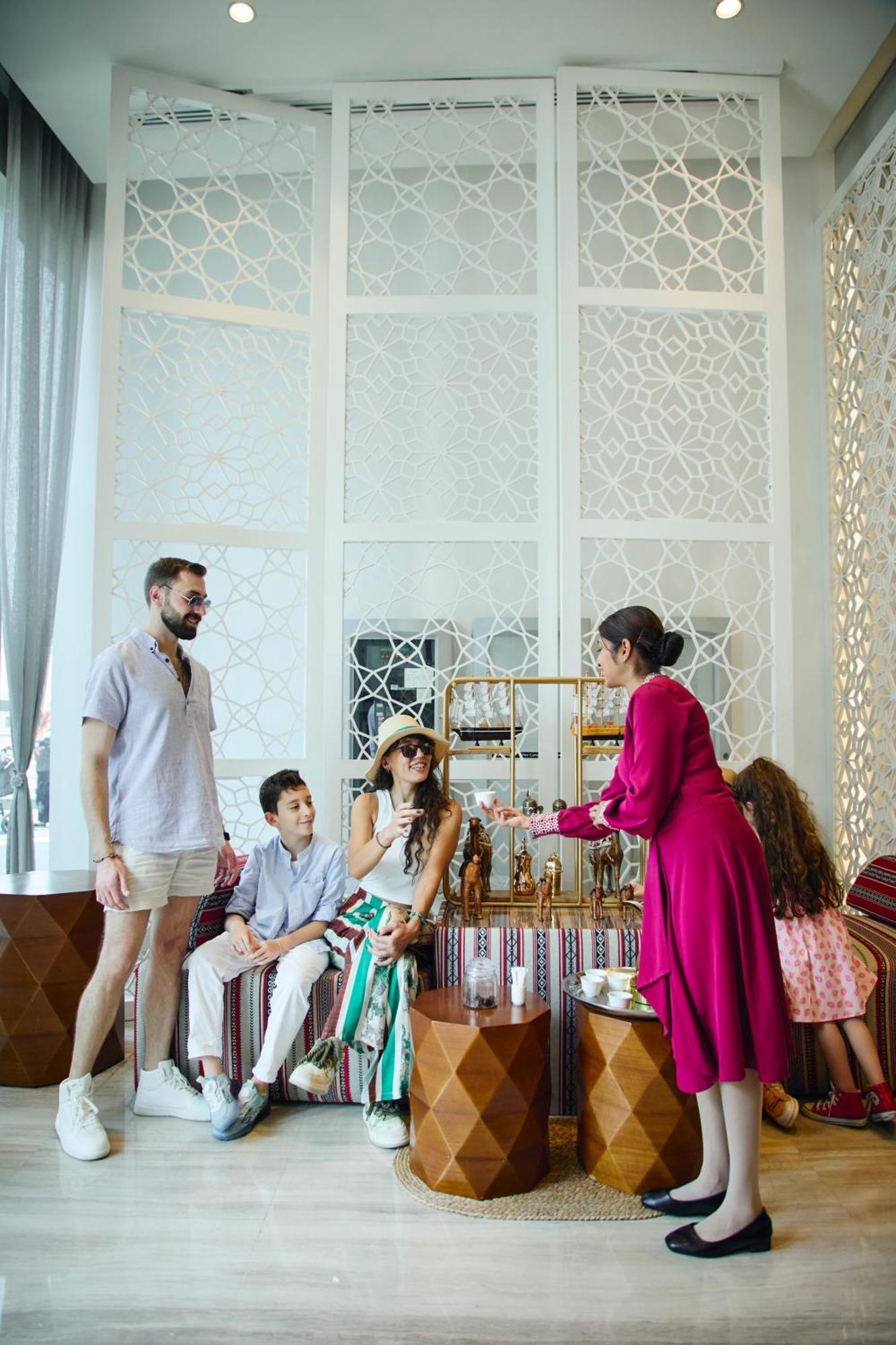 Grand Mercure Dubai City Hotel Exterior photo The photo depicts a social setting where two adults and two children are engaged in a relaxed conversation. One adult is standing while the other is seated, wearing sunglasses and a large hat, suggesting a casual and stylish atmosphere. The seated in