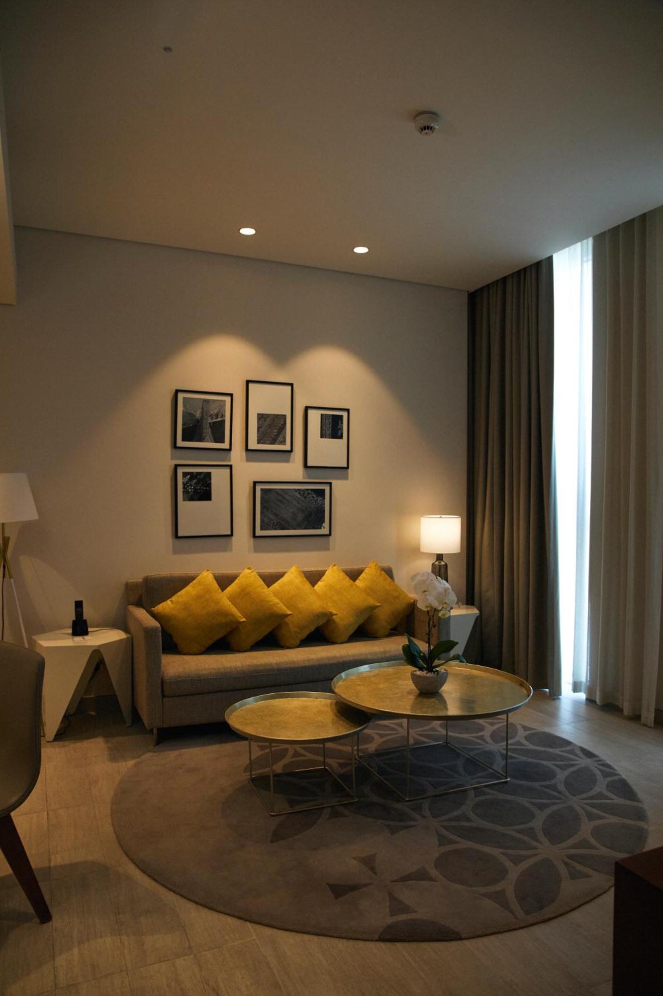 Grand Mercure Dubai City Hotel Exterior photo The photo shows a modern living room setting. There's a light gray sofa adorned with several yellow decorative pillows. Above the sofa, there are four framed artworks hanging on the wall, featuring simple designs. A lamp stands next to the sofa, prov