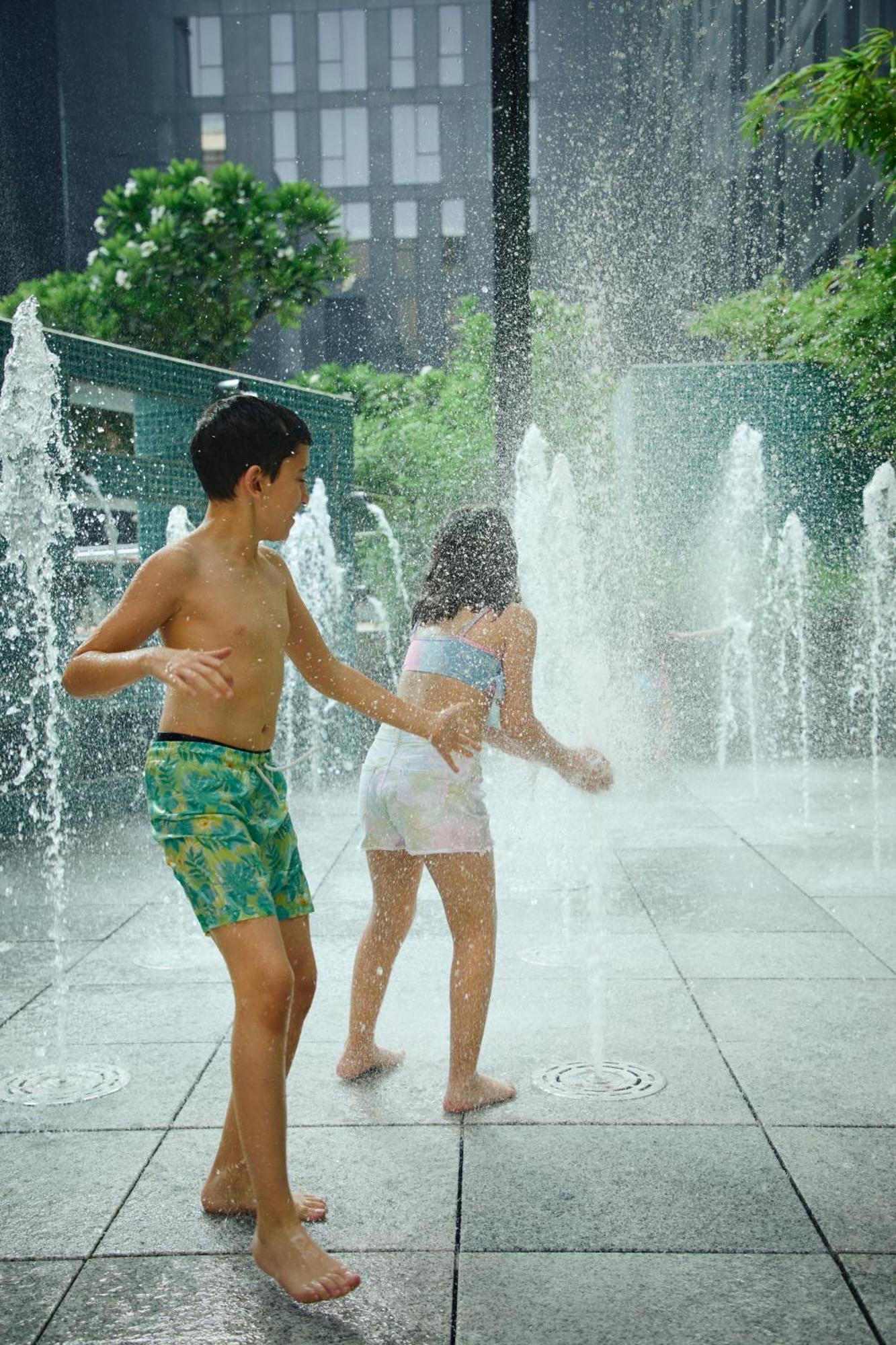 Grand Mercure Dubai City Hotel Exterior photo The photo shows two children playing in a water fountain. The boy, wearing colorful swim trunks, is turned sideways with his arm outstretched. The girl, in a light-colored bathing suit, is seen from behind as she interacts with the water. There are s