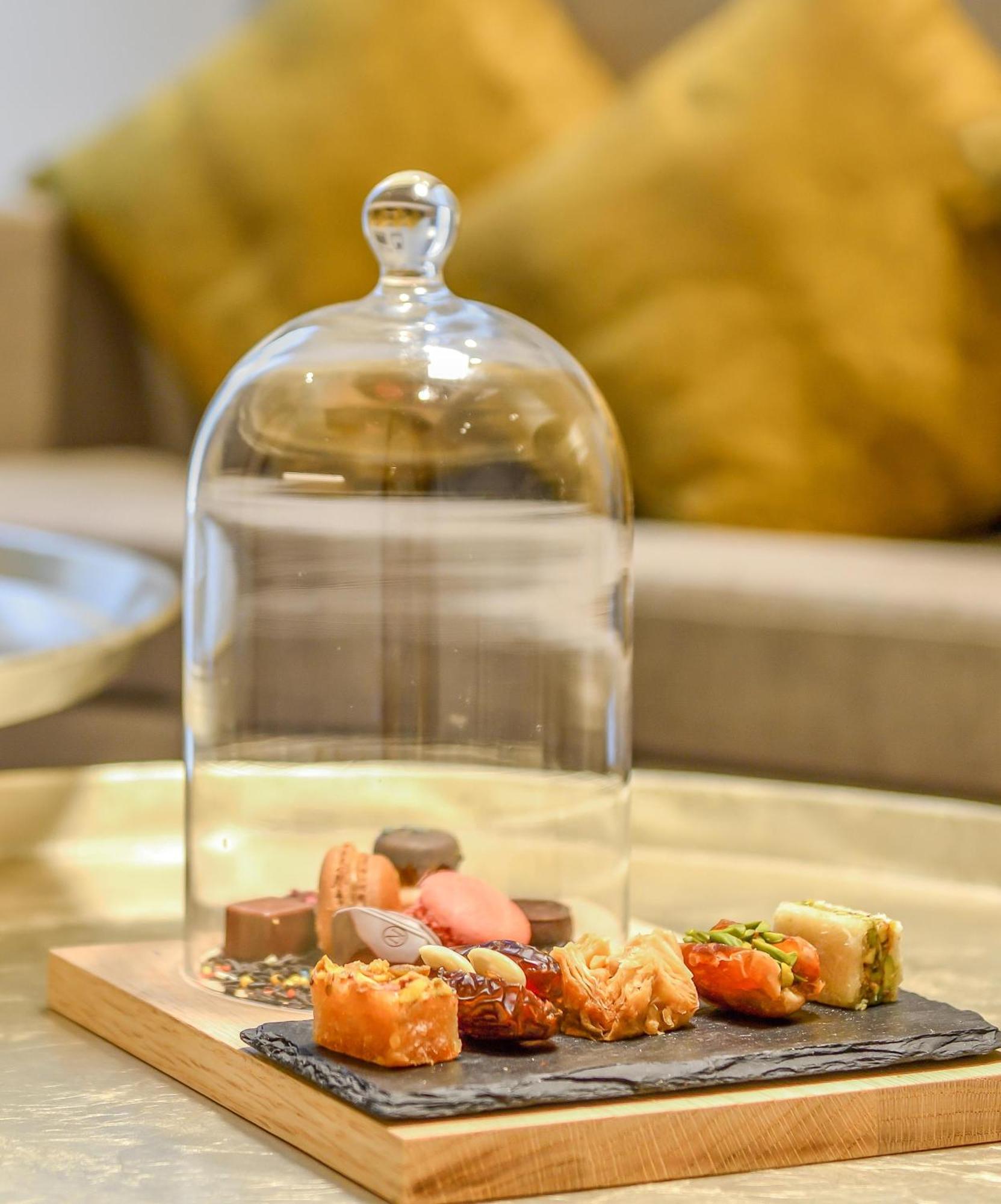 Grand Mercure Dubai City Hotel Exterior photo The photo shows a glass cloche placed over an assortment of assorted pastries and sweets displayed on a wooden board. The treats include colorful macarons and various small desserts, arranged neatly. The background features a soft, blurred couch and 