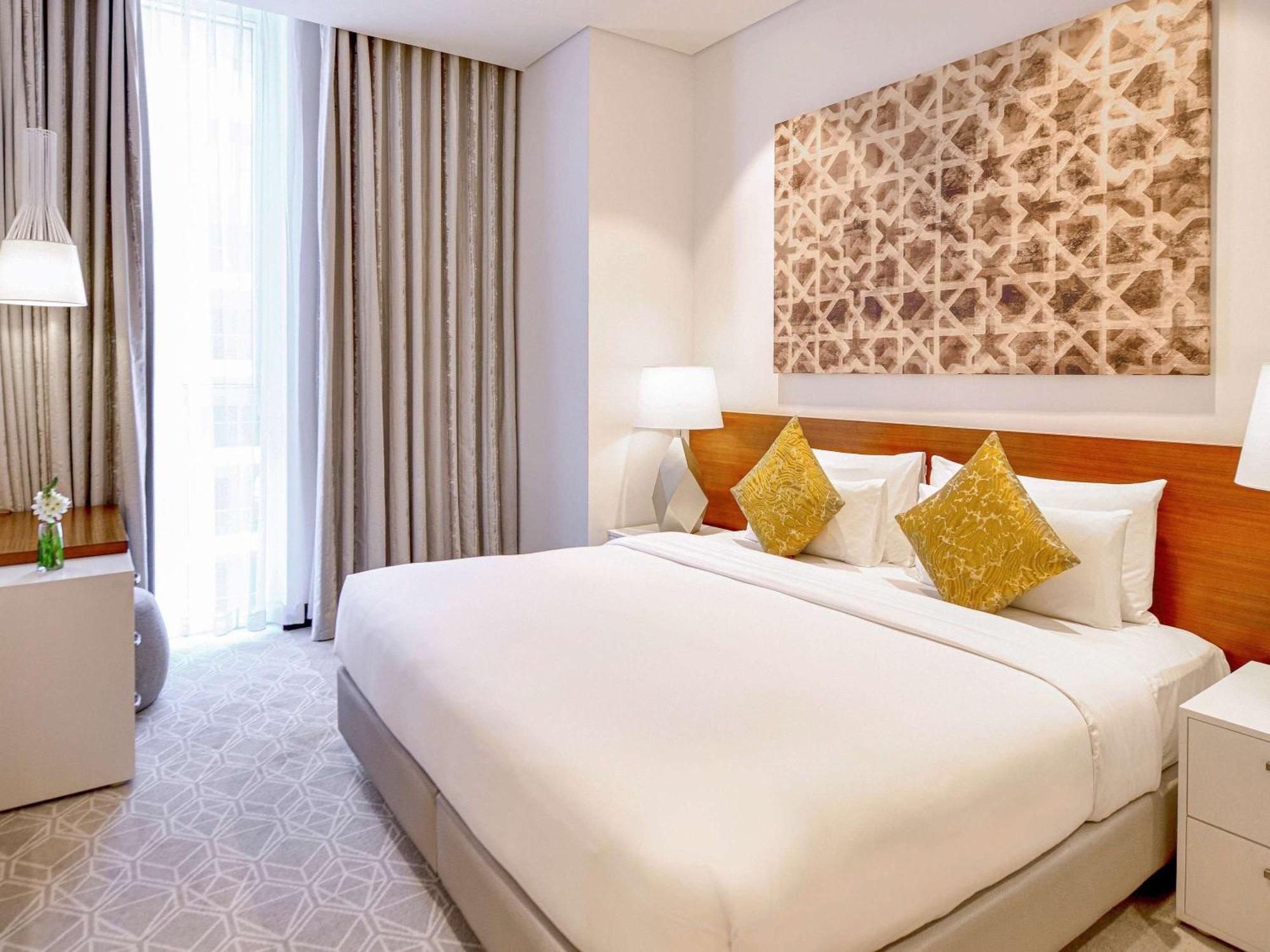 Grand Mercure Dubai City Hotel Exterior photo The photo shows a modern hotel room featuring a large bed with white linens and decorative yellow pillows. There are bedside tables on either side of the bed with lamps. The room has large windows with light-colored curtains, allowing natural light t