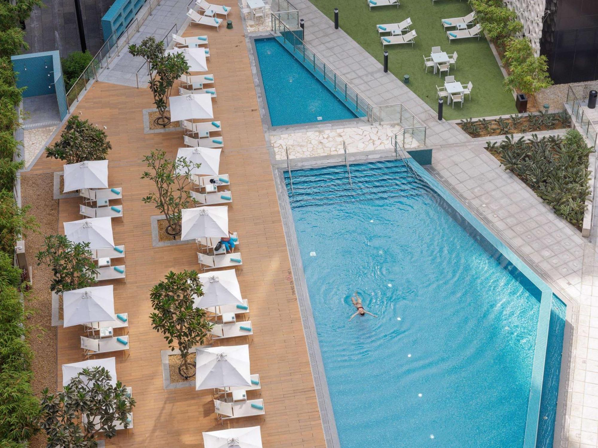 Grand Mercure Dubai City Hotel Exterior photo The photo shows a modern outdoor swimming area with two pools. One pool is larger, featuring a swimmer, while the other is smaller and narrower. Surrounding the pools are several lounge chairs with white umbrellas, providing shaded areas for relaxati